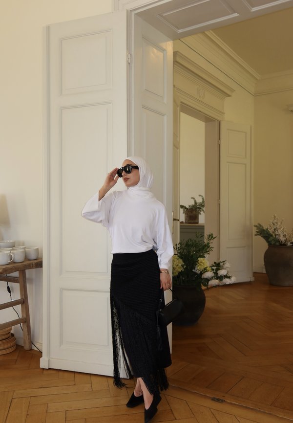 Frau in weißem Hijab und Top mit schwarzem Rock und Sonnenbrille steht in einem Zimmer mit Holzboden und offener weißer Tür.