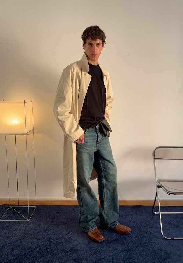 Un hombre luce una gabardina color crema, una camisa negra, unos vaqueros holgados, guantes y zapatos marrones en una habitación minimalista, junto a una lámpara de pie encendida.