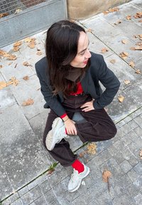 Μαύρο σακάκι πάνω από κόκκινο μπλουζάκι, καφέ παντελόνι από χοντρή βελόνα και ανοιχτό γκρι αθλητικά παπούτσια. Το φόντο περιλαμβάνει τσιμεντένια πλακάκια και πεσμένα φύλλα.