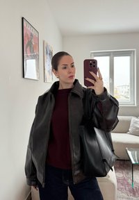 Giacca di pelle nera con colletto alto, maglione bordeaux sotto e borsa tote nera. Tessuto leggermente strutturato e design contemporaneo.