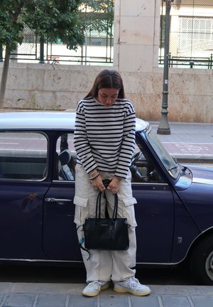 Bolso negro sostenido por una persona que lleva una camisa de rayas de manga larga, pantalones cargo beige y zapatillas, de pie junto a un coche clásico azul.