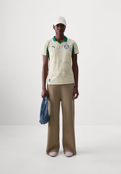 Camisa Puma en un amarillo pálido con acentos verdes, con un diseño texturizado, y una gorra a juego. Combinada con pantalones de pierna ancha beige y calzado rosa.