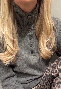 Pull gris côtelé avec un col montant, doté de cinq boutons noirs à l'avant. Pantalon à motifs visibles, présentant un design imprimé léopard.