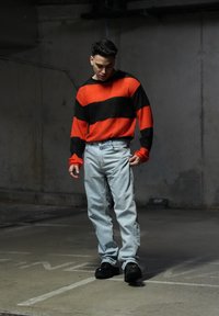 Κόκκινος και μαύρος ριγέ πουλόβερ, ανοιχτό μπλε τζιν και μαύρα χοντρά παπούτσια. Ο πουλόβερ έχει μακριά μανίκια και άνετη εφαρμογή. Σκηνικό από σκυρόδεμα.