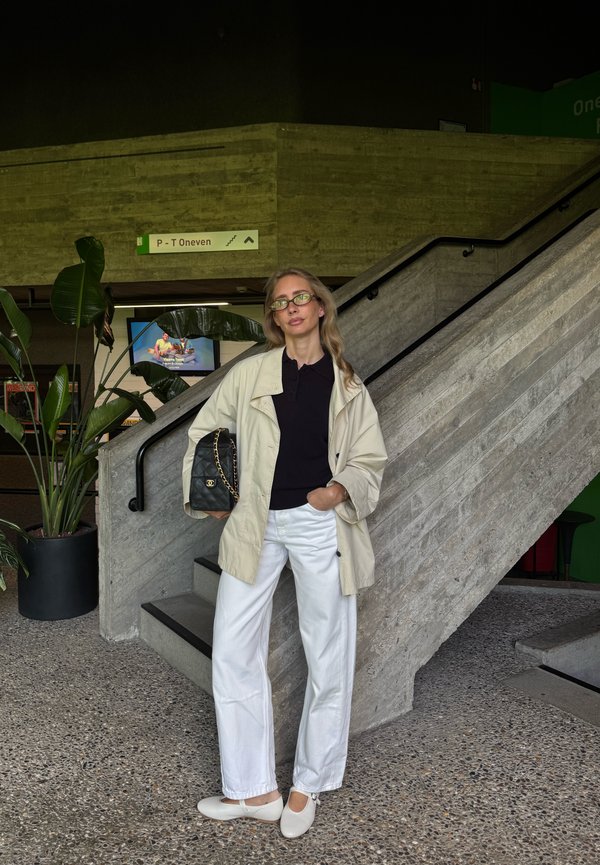 Femme avec des lunettes portant une veste beige et un pantalon blanc debout près d'un escalier en béton tenant un sac à main noir à l'intérieur.