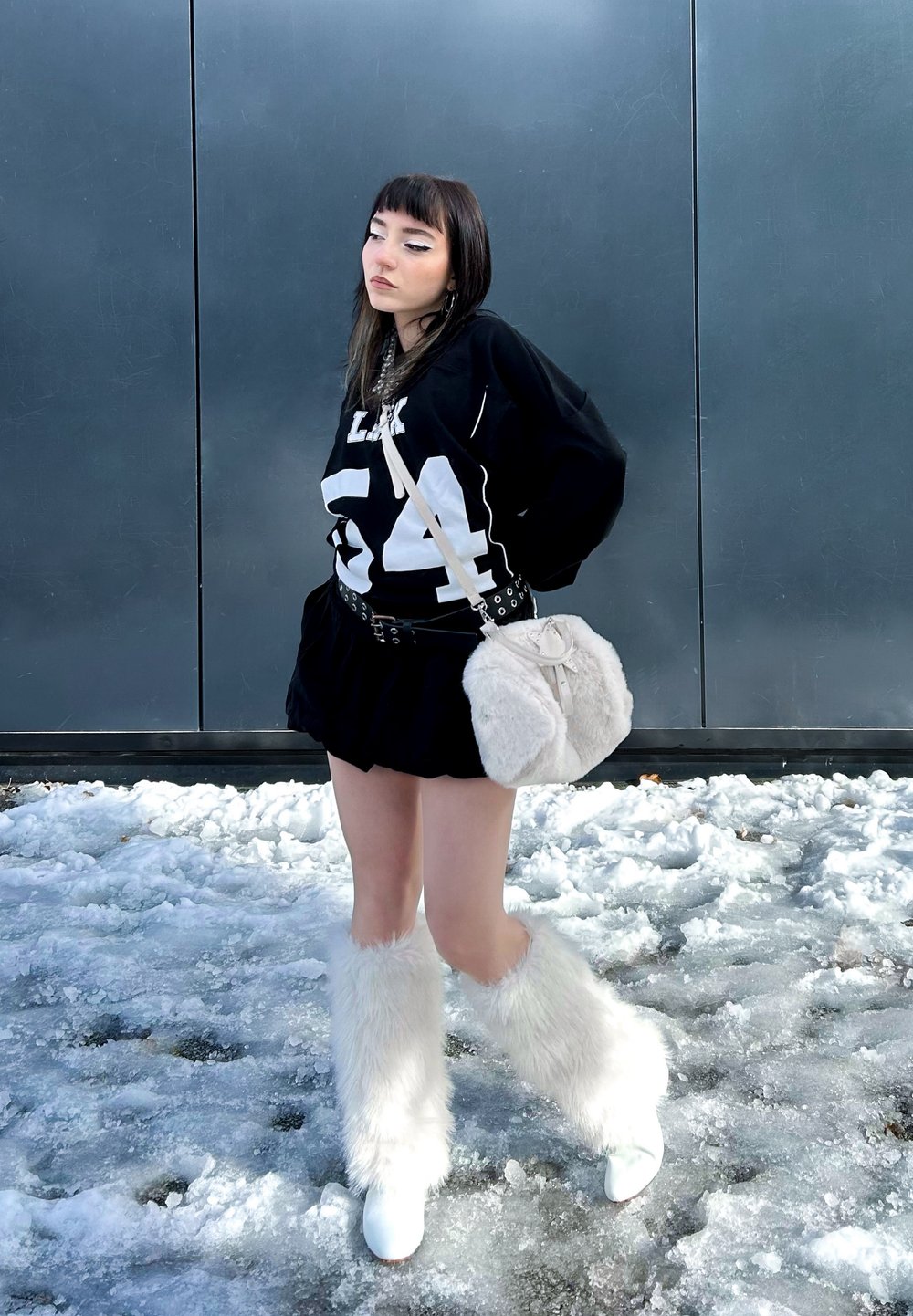 Sweatshirt oversized preto com o número branco "54", saia preta, manoplas brancas peludas, botas brancas de salto e uma mala de pelo creme. Chão coberto de neve.