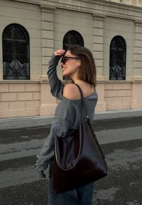 Donna con maglione grigio scollato e occhiali da sole, che porta una grande borsa di pelle marrone scuro, camminando per una strada cittadina con finestre decorative.
