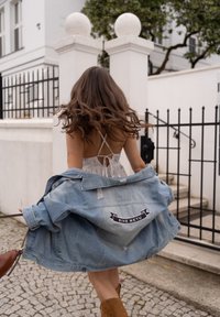 Casaco de denim com detalhes desgastados, apresentando um patch em preto e branco "SINE METU". Usado sobre um vestido claro com padrão floral e botas castanhas.
