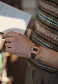 Orologio digitale da polso con quadrante rettangolare di colore bronzo, cinturino in acciaio e display LCD. Chi lo indossa tiene in mano un libro aperto, indossando un maglione con motivo a fantasia.