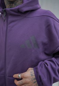 Lila zip-up-huvtröja av mjukt material med en svart logotyp med tre diagonala ränder på vänster sida.
