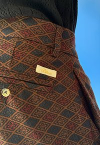 Brun mønstret stoff med diamantdesign og røde detaljer, en knapp, og en liten etikett sydd inn i lommen.