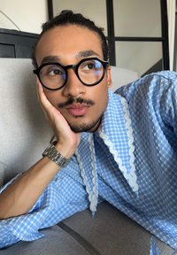 Blå gingham skjorte med dekorativ hvid bølgede kant, lange ærmer og en afslappet pasform. Person med sølvur hviler hagen på hånden.