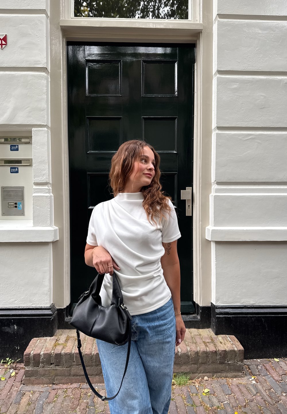 Hvit kortermet topp med drapert design, kombinert med lyseblå jeans og en svart skinnveske, foran en grønn dør og mursteinssti.