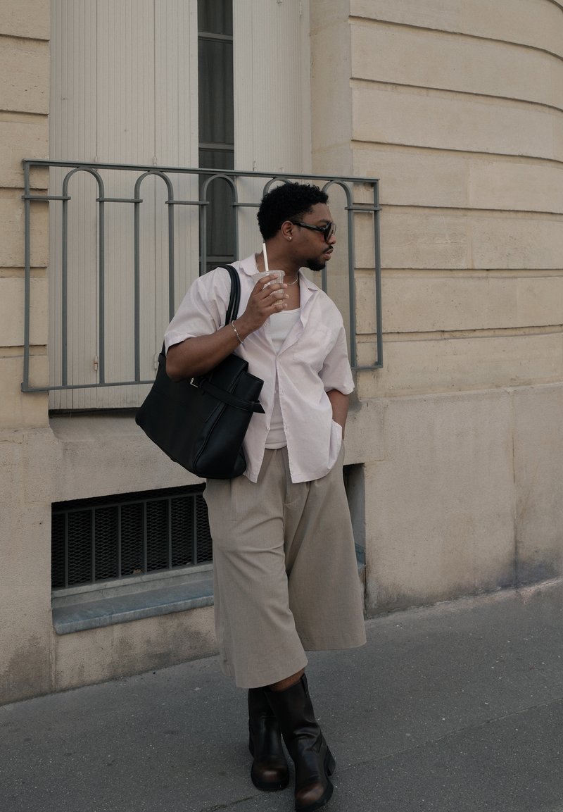 Homme portant des lunettes de soleil et des vêtements décontractés, tenant une boisson et un sac fourre-tout noir, debout devant un bâtiment en pierre beige sur un trottoir.