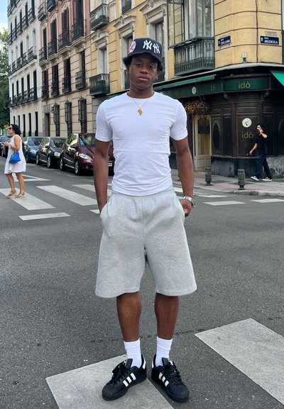 Camiseta blanca, pantalones cortos grises, zapatillas negras con rayas blancas. De pie en un paso de peatones, fondo urbano con edificios y coches estacionados.