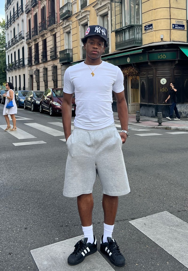 T-shirt blanc, shorts gris, baskets noires avec des rayures blanches. Debout sur un passage piéton, arrière-plan urbain avec des bâtiments et des voitures garées.