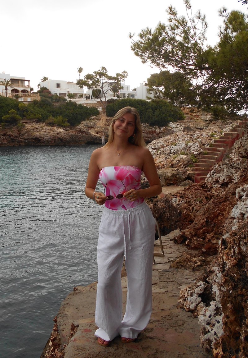 Young woman in white trousers and a pink floral top, holding sunglasses, standing on a rocky coast near calm water and some stairs.