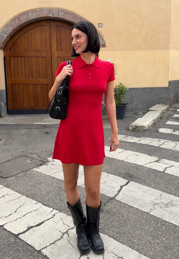 Femme avec coiffure au carré noir portant une robe rouge à manches courtes et des bottes noires, debout sur un passage pour piétons près d'une porte en bois cintrée.