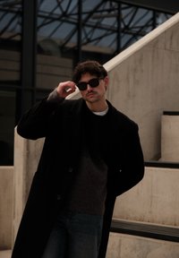 Homme aux cheveux bouclés portant des lunettes de soleil, un manteau noir et un pull foncé, debout près d'un escalier en béton et d'un bâtiment vitré.