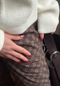 Mains aux ongles rouge foncé reposant sur un tissu marron à motifs, portant un pull en maille blanche et tenant une ceinture en cuir marron foncé avec une boucle en argent.