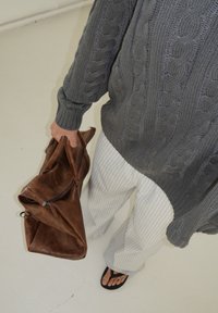 Suéter cinza de malha trançada, calças largas listradas em bege, mala de couro marrom e chinelos pretos, vistos de cima.