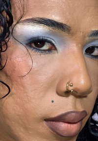 Nærbilde av et ansikt med middels brun hudfarge, som bærer blå øyenskygge, bronsefarget leppestift og gull neseringer, med teksturert hud og markerte bryn.