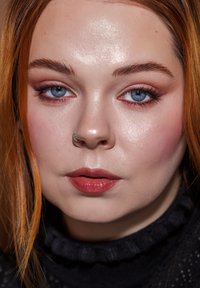 Nærbilde av et ansikt med glatt hud, klare blå øyne, lett sminke, kobberfarget hår og en sølvfarget hoops-nese ring, iført en svart teksturert genser.