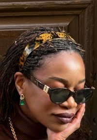 Femme aux cheveux tressés portant des lunettes de soleil écaille de tortue, un bandeau à imprimé léopard, des boucles d'oreilles pendantes vertes et un rouge à lèvres foncé, reposant son visage sur sa main.