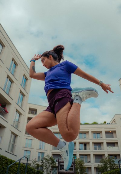 Calzado deportivo en azul claro con detalles en blanco, combinado con shorts morados y una camiseta azul, en una pose de salto en el aire con un fondo de edificio.