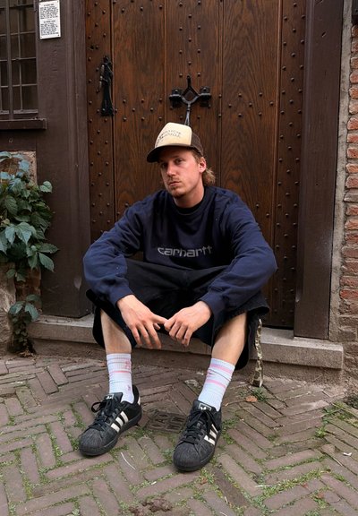 Joven con gorra y sudadera azul marino sentado en un escalón de piedra frente a una gran puerta de madera con tachuelas de metal.