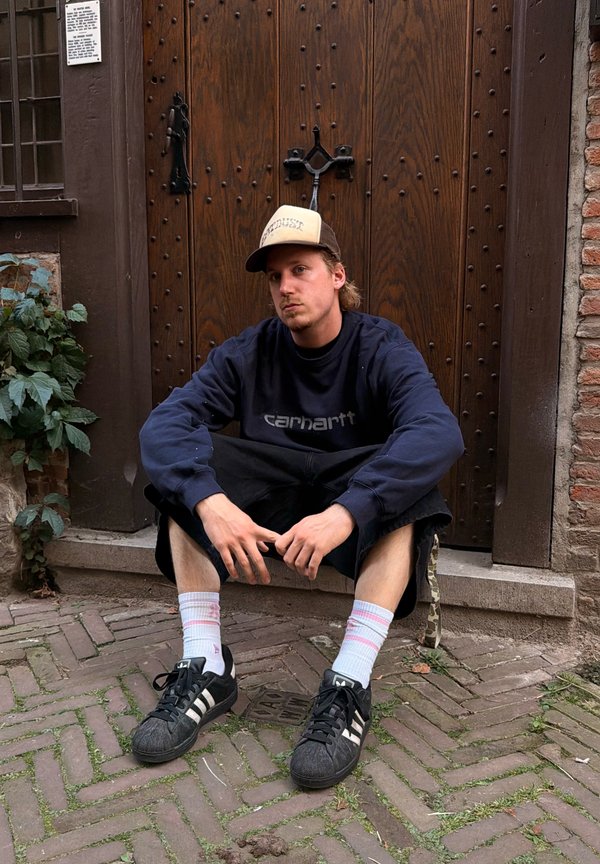 Jeune homme portant une casquette et un sweat-shirt bleu marine assis sur une marche en pierre devant une grande porte en bois avec des clous en métal.