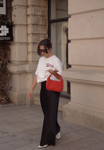 Camiseta blanca con diseño gráfico, pantalones anchos negros, bolso acolchado rojo y zapatos crema. Gafas de sol y teléfono en la mano. Ambiente urbano.