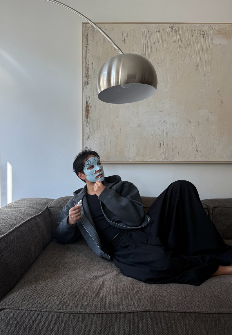 Personne avec un masque facial bleu appliquant un soin de la peau en se penchant sur un canapé gris sous une grande lampe sur pied en arc métallique et une œuvre murale texturée.