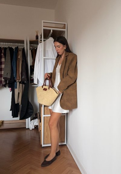Blazer oversize de ante marrón, vestido blanco, zapatos planos marrones y bolso trenzado beige con asas marrones. Muebles de madera clara y perchero en el fondo.