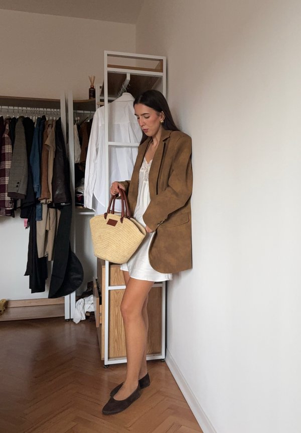 Hnedý semišový oversize blejzer, biele šaty, hnedé baleríny a tkaná svetlohnedá taška s hnedými rukoväťami. V pozadí svetlé drevené nábytkové vybavenie a stojan na oblečenie.
