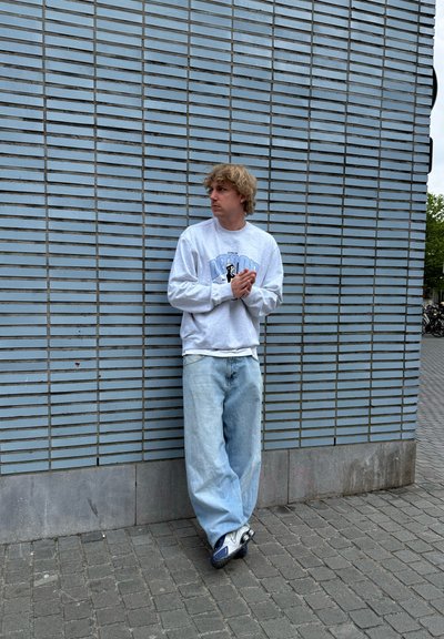 Sudadera gris claro con diseño gráfico, jeans sueltos azul claro y zapatillas negras. De pie contra una pared de ladrillos azules texturizados.