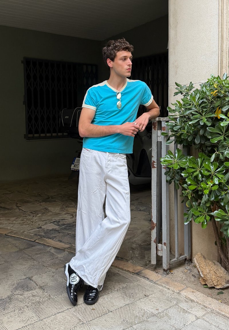 Jeune homme en chemise turquoise et pantalon blanc appuyé contre une porte à côté d'un arbuste vert dans un espace extérieur ombragé avec un sol en pierre à motifs.