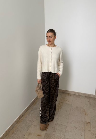 Mujer de pie en el interior con un cárdigan blanco abotonado, pantalones sueltos con estampado de leopardo, zapatos marrones claros y sosteniendo un bolso beige.