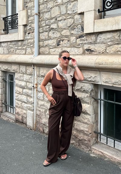 Top marrón sin mangas combinado con pantalones marrones de cintura alta, un suéter beige claro colgado sobre los hombros y gafas de sol negras.