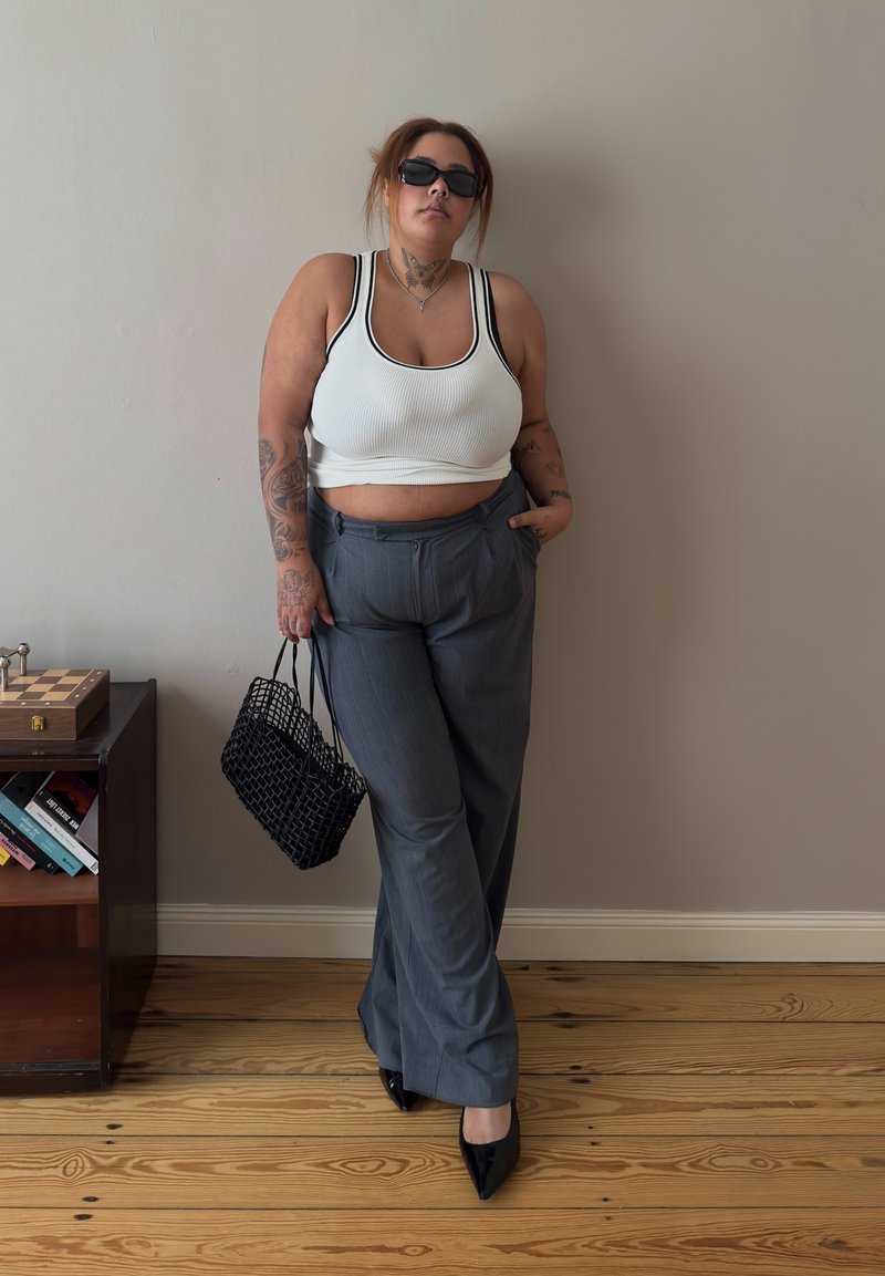 Donna con tatuaggi che indossa occhiali da sole, top corto bianco, pantaloni grigi, tacchi neri, tiene una borsa nera a rete, in piedi contro una parete beige accanto a una mensola di legno.