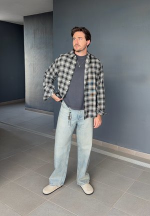 Hombre con barba de pie, vistiendo camisa de cuadros gris sobre camiseta oscura, vaqueros azul claro, zapatos beige, con llaves colgando y mirando de lado en el interior.
