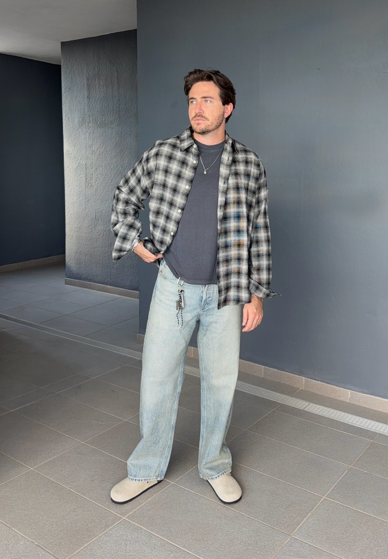 Man met baard staand, draagt een grijze geruite blouse over een donker T-shirt, lichtblauwe spijkerbroek, beige schoenen, met sleutels hangend en kijkt zijwaarts binnen.