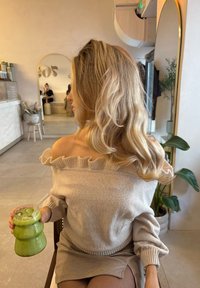 Sweater bege com decote ombro a ombro e gola com babados; servindo uma bebida verde num copo texturizado; saia xadrez; ambiente interno.