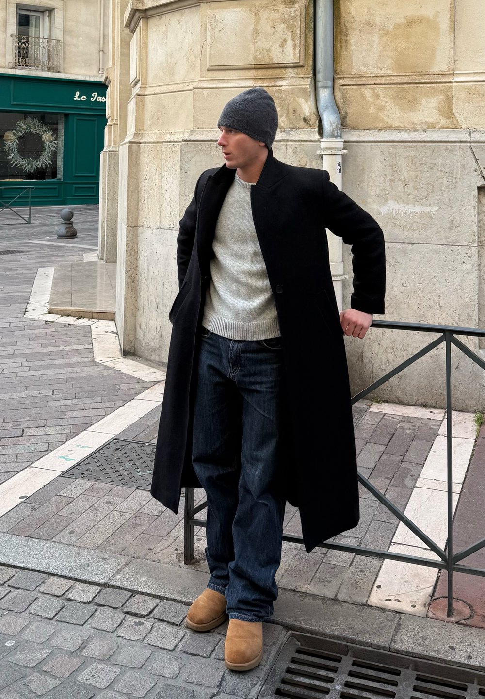Jeune homme portant un bonnet gris, un long manteau noir, un pull léger, un jean et des bottes couleur sable, appuyé sur une rambarde métallique sur un trottoir en ville.