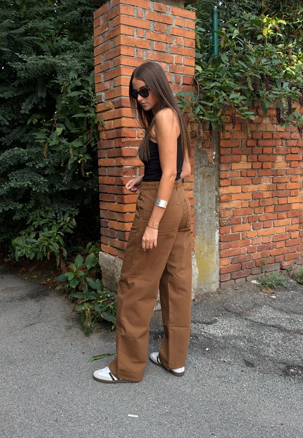 Joven mujer con cabello largo que lleva gafas de sol, una camiseta sin mangas negra, pantalones anchos marrones y zapatos blancos, está de pie junto a una pared de ladrillos al aire libre.