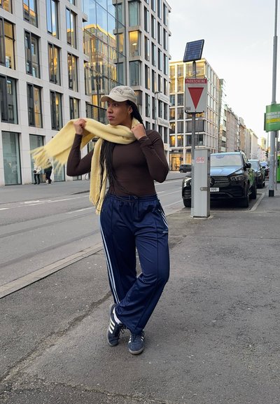 Una persona lleva una camiseta de manga larga marrón, pantalones deportivos azul marino con rayas blancas y una bufanda de color amarillo claro. Está sosteniendo la bufanda.