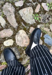 Mocassins de couro preto com detalhes em costura, usados com meias brancas e calças pretas listradas, sobre uma superfície de pedra texturizada.