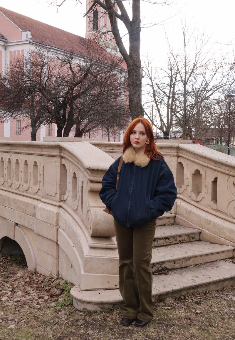 Jeune femme aux cheveux roux portant une veste sombre à col en fourrure, debout près d'un escalier en pierre à l'extérieur d'un bâtiment, entourée d'arbres sans feuilles en hiver.