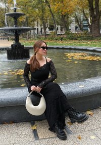 Haut en dentelle noir à manches longues et jupe, bottes noires à semelle plateforme et lacets, tenant un sac à main blanc courbé, assise sur un banc en pierre près d'une fontaine.