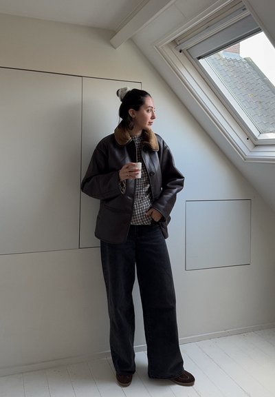 Chaqueta de cuero marrón con un cuello de piel marrón, combinada con una camisa de cuadros y jeans oscuros holgados. De pie junto a una claraboya en una habitación luminosa.